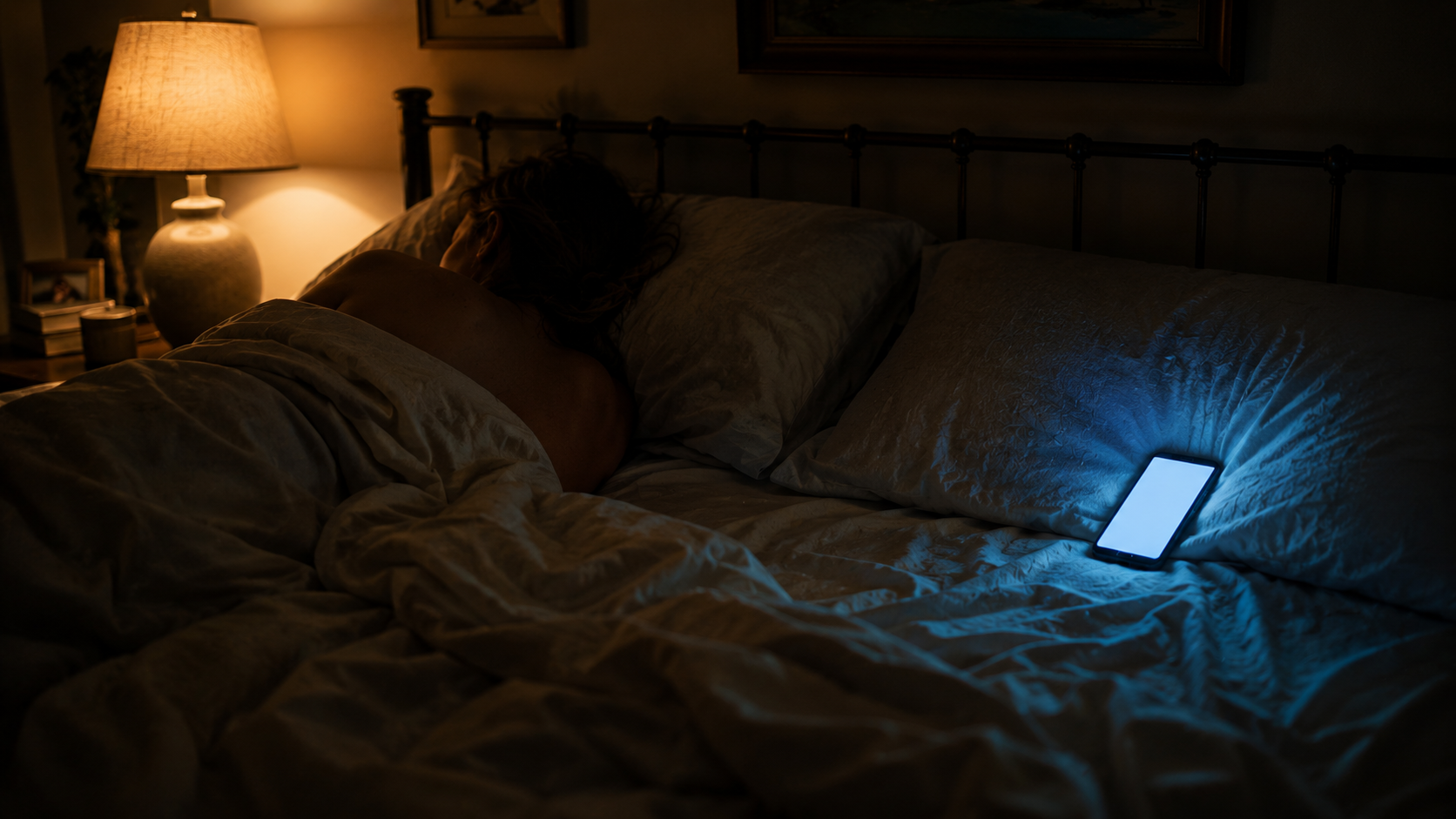 A bedroom at night — one person asleep, turned away on the lamp-lit side; a phone glowing cold blue on the empty second pillow
